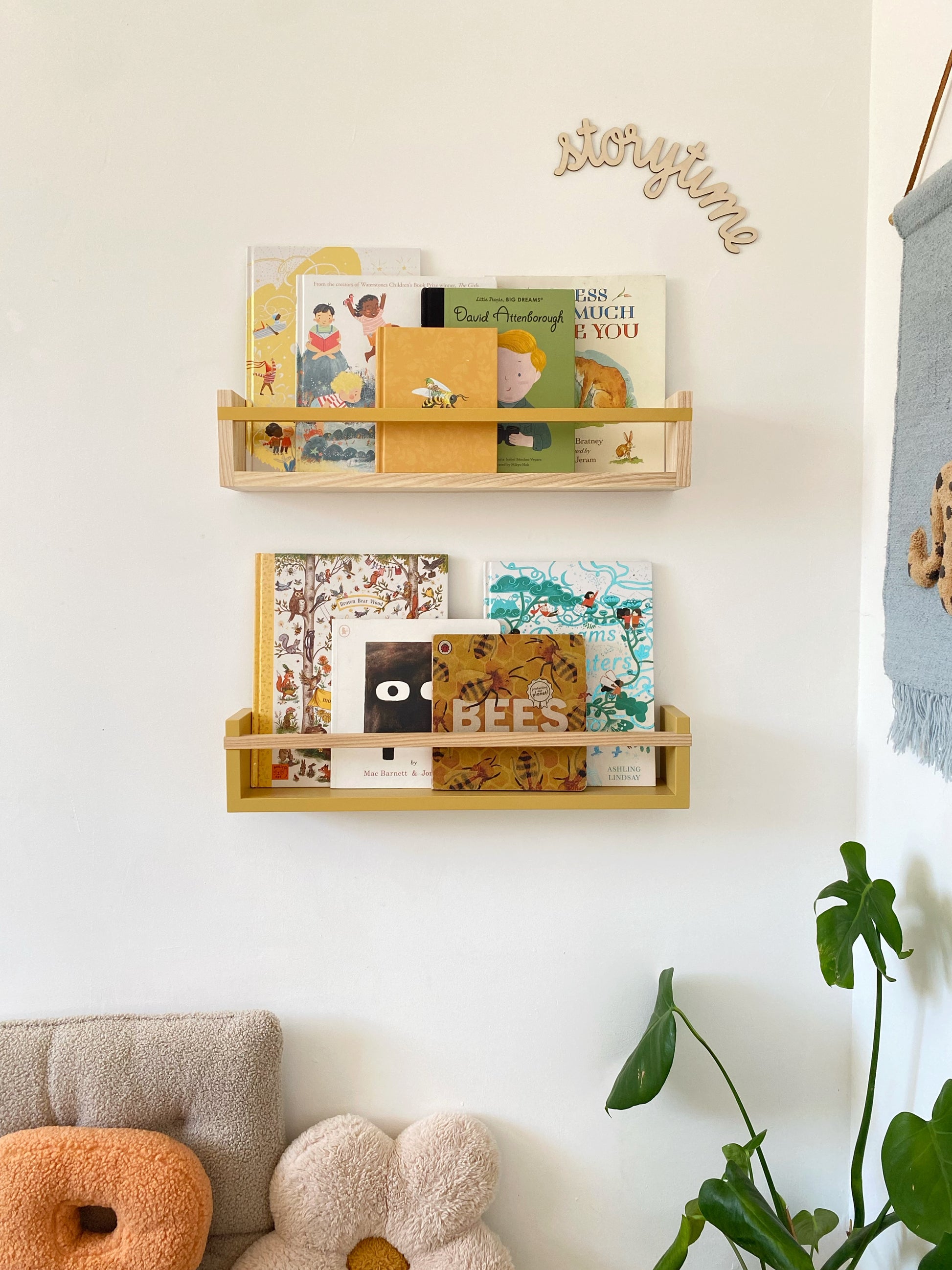 Wooden bookshelf with children's books against a white wall, with a plant and sofa in the foreground.