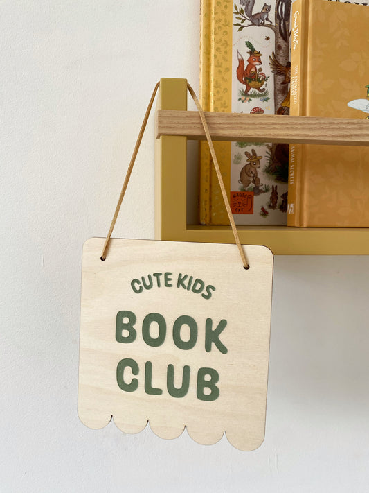 Wooden sign with 'Cute Kids Book Club' text hanging in front of a bookshelf.