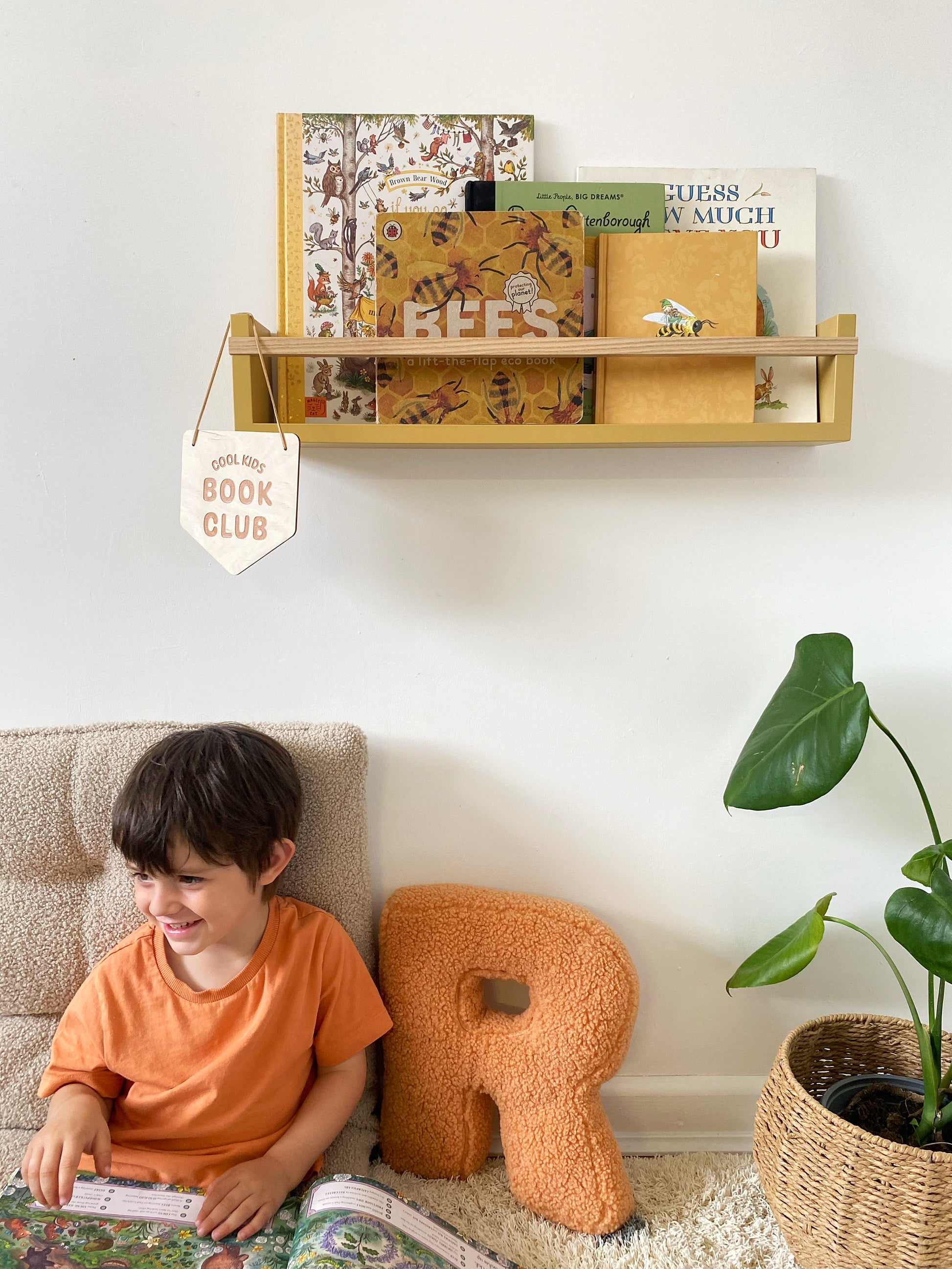 Child sitting on a couch with a book, next to a shelf with books and a plant, in a room setting.