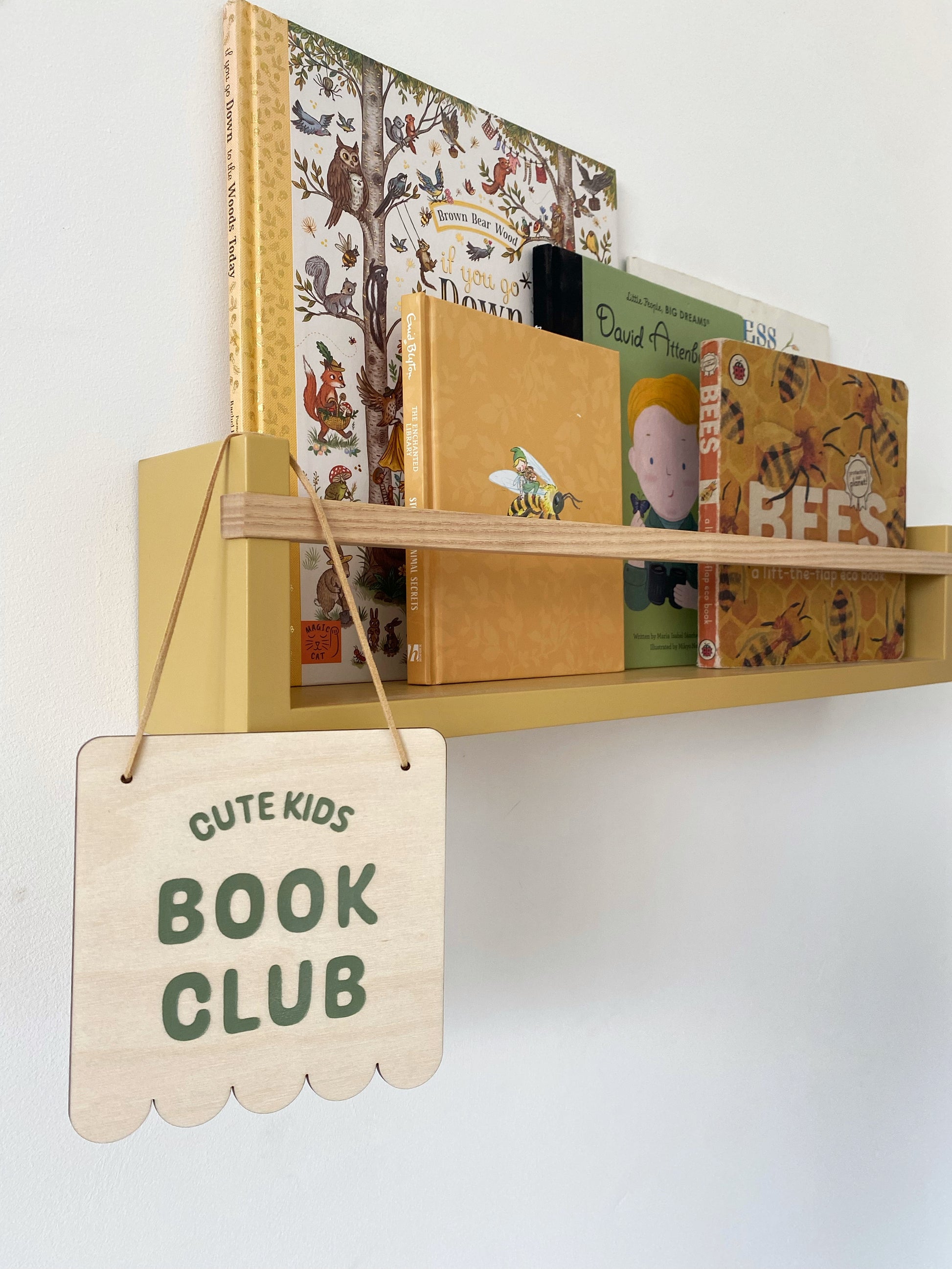 Shelf with children's books and a sign that reads 'Cute Kids Book Club' on a white wall.
