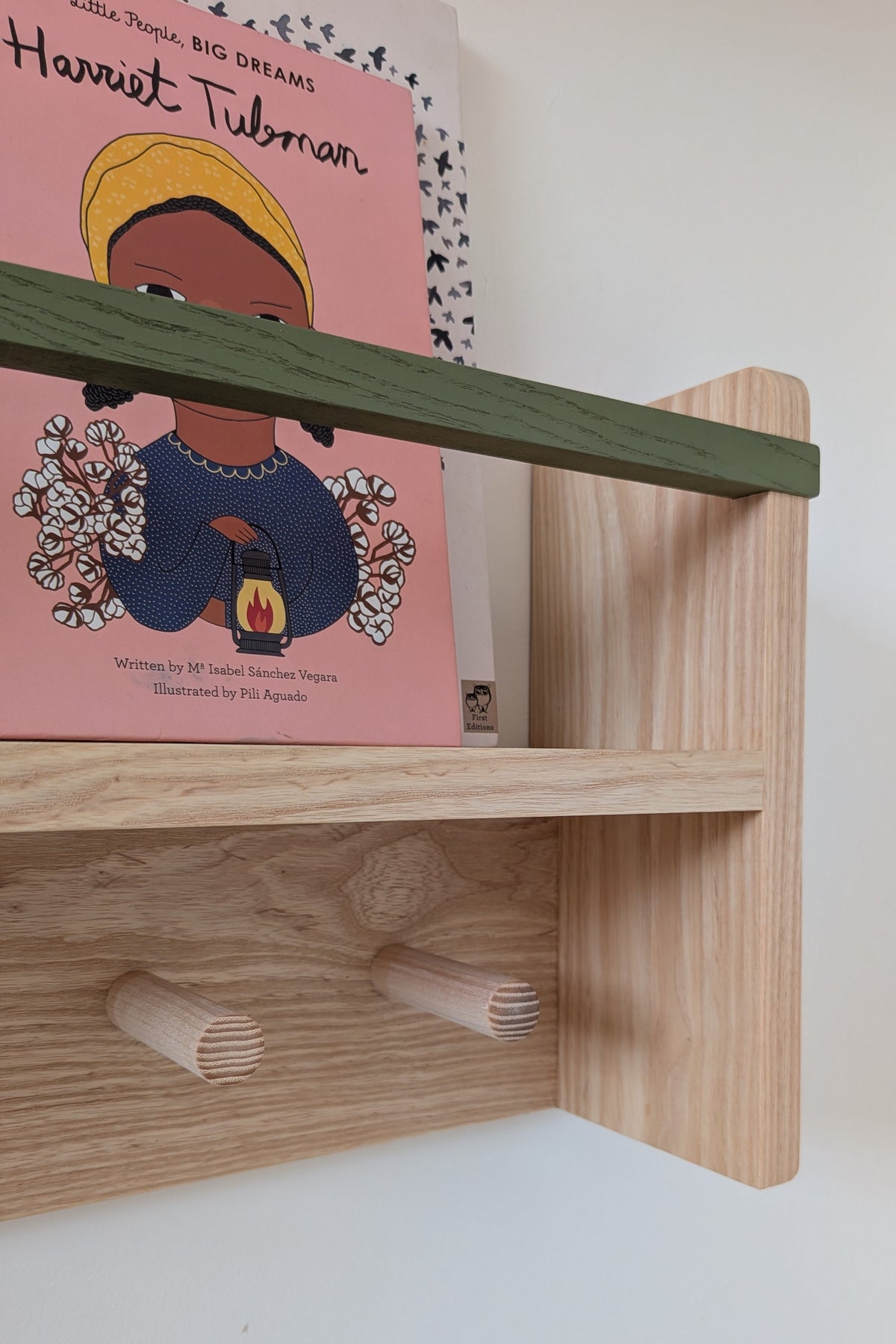 A light natural wood bookshelf with a green-painted top rail holding a children's book titled 'Harvest Tubman' against a white wall.