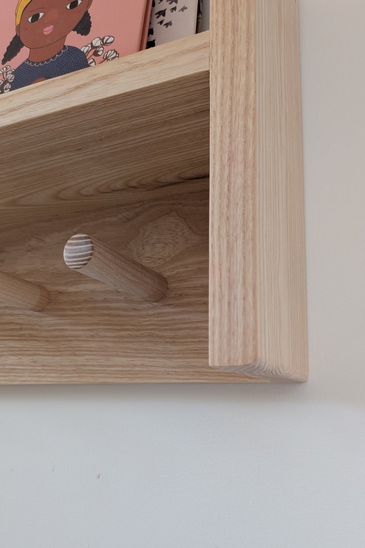 Close-up of a light natural wood wall-mounted shelf with wooden peg hooks, shown against a plain off-white wall with a small portion of a colorful book visible at the top.