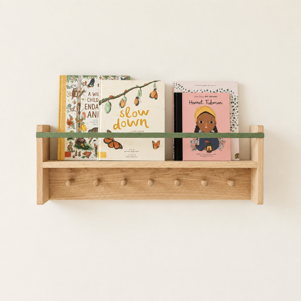Two light natural wood wall-mounted bookshelves on a warm off-white wall, styled with children's books. The lower shelf has a green glass shelf rod and peg hooks, while the upper shelf has a pink wooden rod.
