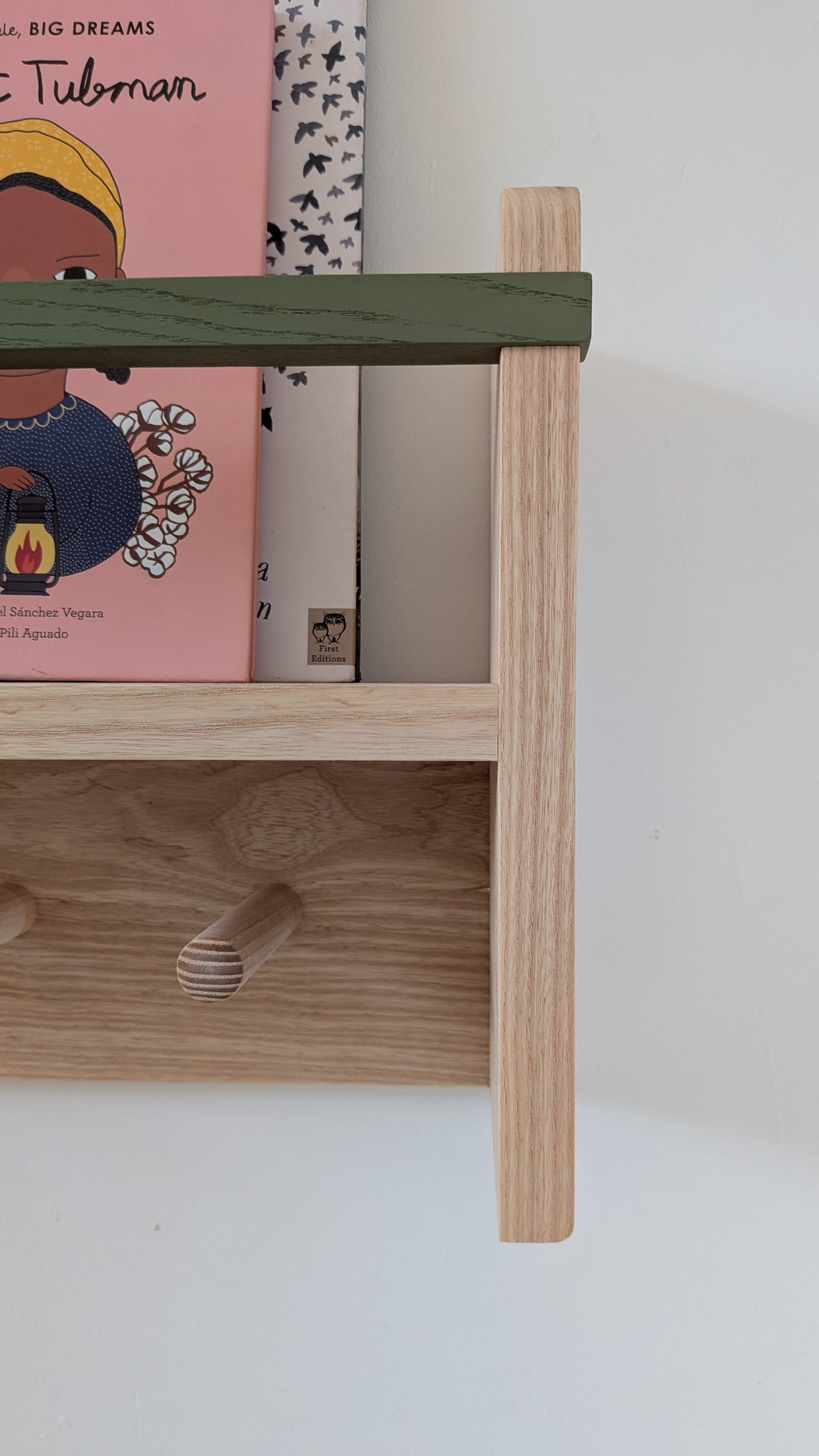 A close-up of a light natural wood bookshelf with a dark green horizontal bar, holding children's books against a plain white wall.