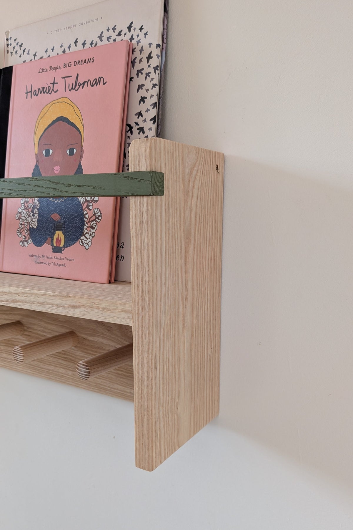 A light natural wood floating bookshelf mounted on a white wall, holding a pink children's book and a black-and-white patterned book, with a green horizontal bar across the shelf.